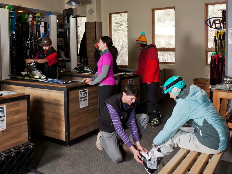 Inside the rental shop: staff help guests with skis and boots at wooden counters in front of racks of rental skis
